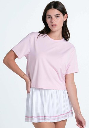 Jonge vrouw met donker haar die een lichtroze T-shirt met korte mouwen en een witte geplooide rok met roze strepen draagt, staand tegen een effen achtergrond.