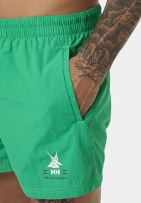 Zelené plavkové šortky s páskem na stažení, bočními kapsami a bílým logem se stylizovanou plachetnicí a názvem značky na přední straně.