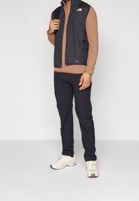 Gilet leggero nero sopra una maglietta a maniche lunghe marrone, abbinato a pantaloni cargo neri e scarpe da ginnastica bianche, dotato di cerniere e tasche.