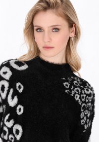 Schwarzer Pullover mit flauschiger Textur, der auf einem Ärmel und der Schulter weiße Leopardenmuster-Akzente aufweist. Rundhalsdesign.