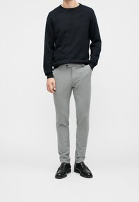 Donkerblauwe trui met ronde hals gecombineerd met lichtgrijze, aansluitende broek. Schoenen zijn zwarte loafers. Soepele textuur en een nauwkeurige, op maat gemaakte uitstraling.