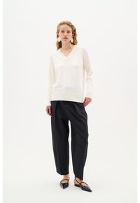 Cremfarbener V-Ausschnitt-Pullover mit geripptem Saum, kombiniert mit lockeren schwarzen Hosen und spitzen schwarzen Ballerinas. Abgebildetes Modell mit schulterlangem, welligem Haar.
