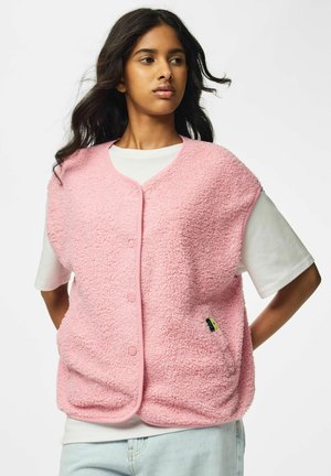 Jeune femme portant un gilet rose duveteux sans manches boutonné par-dessus une chemise blanche à manches courtes, les mains sur les hanches.