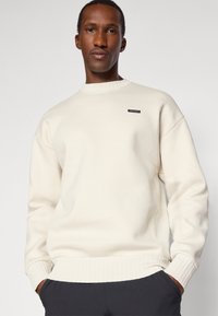 Sweatshirt de couleur crème avec col et poignets côtelés, présentant une petite patch logo noire sur la poitrine. Fabriqué en tissu doux et texturé.