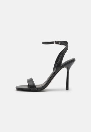 Sandale noire à talon haut avec fine bride à la cheville et boucle, bout ouvert et talon aiguille fin vue de côté sur fond uni.