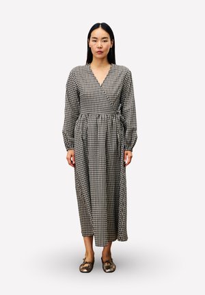 Femme debout portant une robe portefeuille à manches longues à carreaux noir et blanc et des ballerines métalliques sur un fond blanc uni.