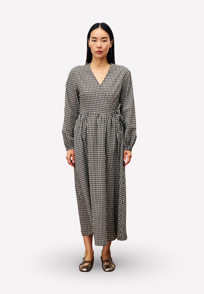 Femme debout portant une robe portefeuille à manches longues à carreaux noir et blanc et des ballerines métalliques sur un fond blanc uni.