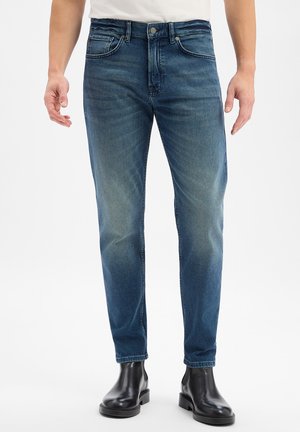 Mann trägt ausgewaschene blaue Slim-Fit-Jeans und schwarze Leder-Stiefeletten vor einem schlichten weißen Hintergrund.