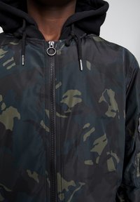 Kurtka camo w ciemnozielonym i czarnym kolorze, z zapięciem na zamek, regulowanym kapturem i teksturowanym materiałem. Posiada srebrne akcenty metalowe.