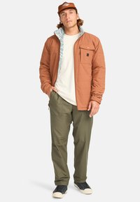 Chaqueta acolchada marrón con un forro azul estampado, bolsillo en el pecho y cuello alto. Pantalones verdes oliva y zapatillas negras de corte bajo.