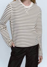 Maglione a righe crema con strisce orizzontali marroni, scollatura a girocollo, polsini a costine e vestibilità morbida, abbinato a pantaloni in denim neri.