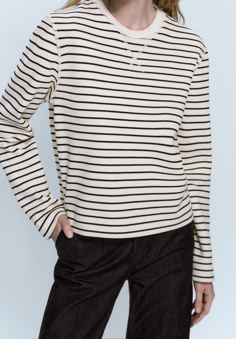 Maglione a righe crema con strisce orizzontali marroni, scollatura a girocollo, polsini a costine e vestibilità morbida, abbinato a pantaloni in denim neri.