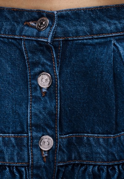 Κοντινό πλάνο ενός σκούρου μπλε denim ενδύματος με τρία εμφανή μεταλλικά κουμπιά και πορτοκαλί ραφές σε μαζεμένο ύφασμα.