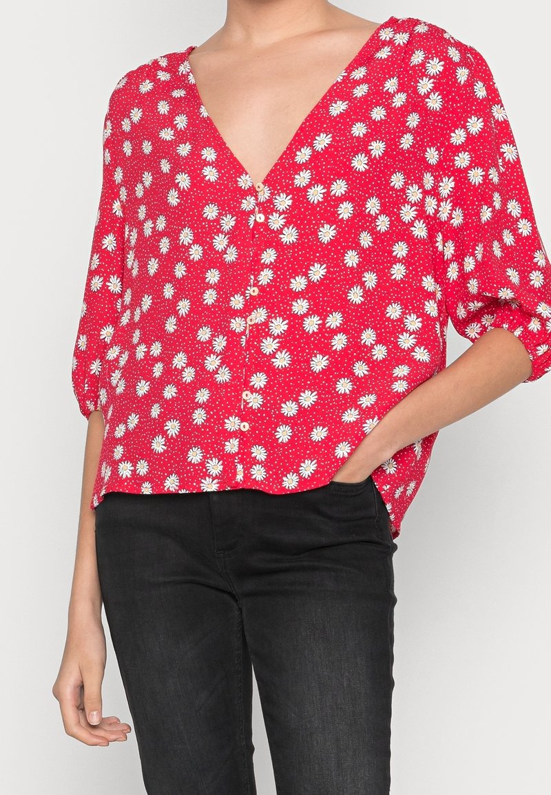 Blouse florale rouge avec des motifs de marguerites blanches, décolleté en V, manches courtes bouffantes et fermeture à boutons à l'avant. Associée à un pantalon noir.