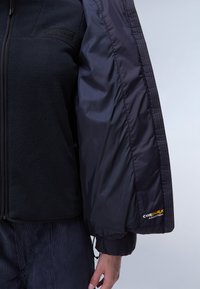 Svart fleecejacka med en slät svart nylonärm. Har Cordura-logotyp på ärmen, med justerbara manschetter och dragkedjestängning.