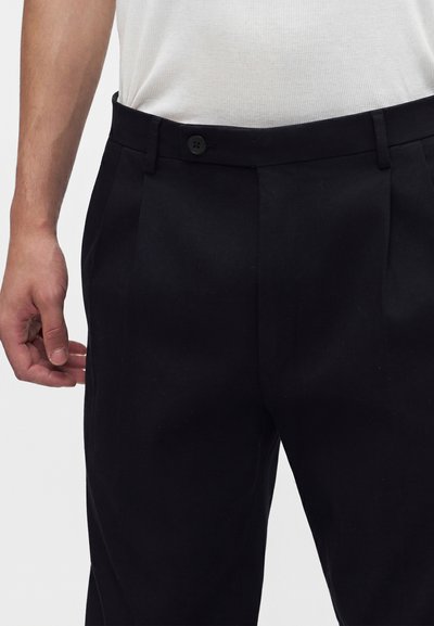 Homme portant un pantalon noir formel avec un bouton et des passants de ceinture, associé à un débardeur blanc côtelé.