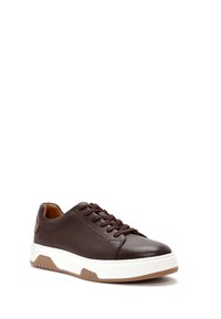 Zapatilla de cuero marrón con lateral texturizado, cordones marrones, suela gruesa blanca y acento tostado en la suela exterior, mostrada en un leve ángulo lateral.
