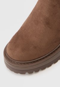 Brown suede ankle boot with rounded toe and textured upper. Thick brown rubber sole with visible tread pattern and stitching detail.