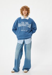 Blauwe sweatshirt met witte tekst "NEW YORK MANHATTAN 1991 UPPER EAST SIDE," gelaagd over een blauw-wit gestreepte colbert, gecombineerd met wijde jeans.