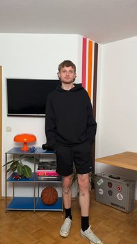 Giovane uomo con felpa nera con cappuccio e pantaloncini si trova in una stanza con una palla da basket, una lampada arancione, una TV, piante e una valigetta argentata con adesivi.
