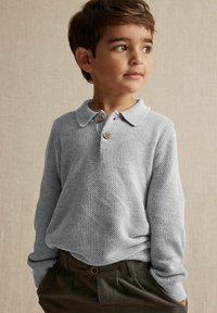 Ljust grått stickat tröja med ett texturerat mönster, krage och tre bruna knappar, tillsammans med mörka, texturerade byxor.