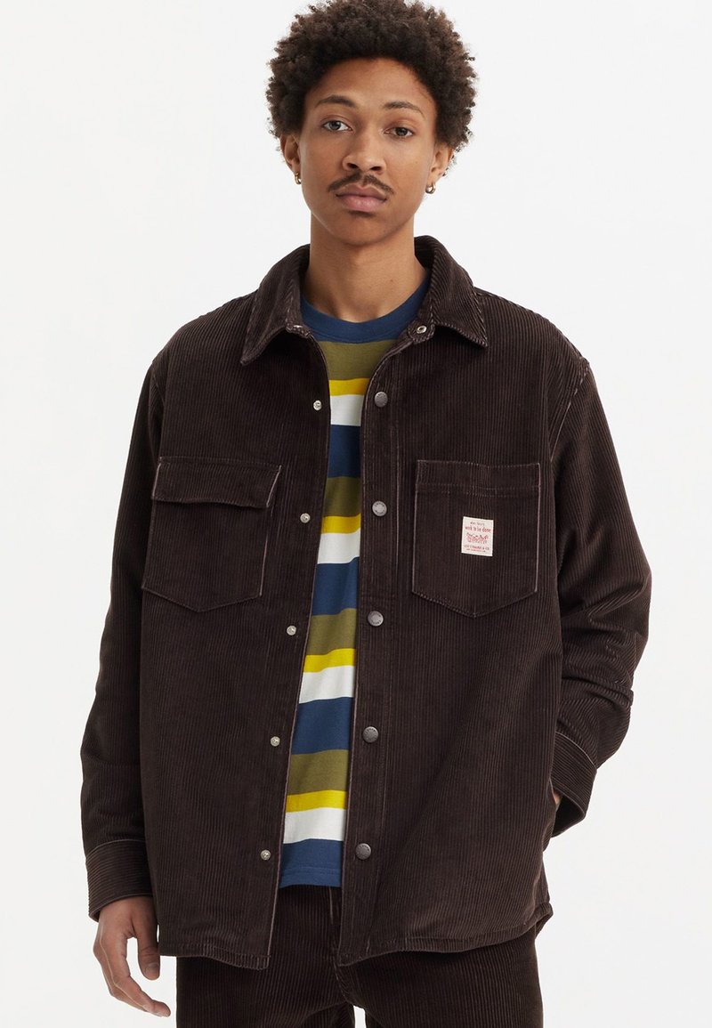 Brown corduroy jacket featuring a button-up front, two chest pockets, and a small branded patch. Dark textured fabric with a relaxed fit.