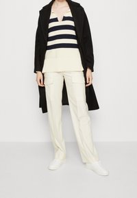 Schwarzer Mantel über einem gestreiften Marineblau- und Creme-Pullover, kombiniert mit cremefarbenen Cargo-Hosen und weißen Sneakers, die einen geschichteten Outfit-Look schaffen.