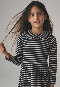 Flicka med långt brunt hår som bär en marinblå och vit randig klänning med rundad krage, håller en hårslinga och tittar lätt åt sidan.