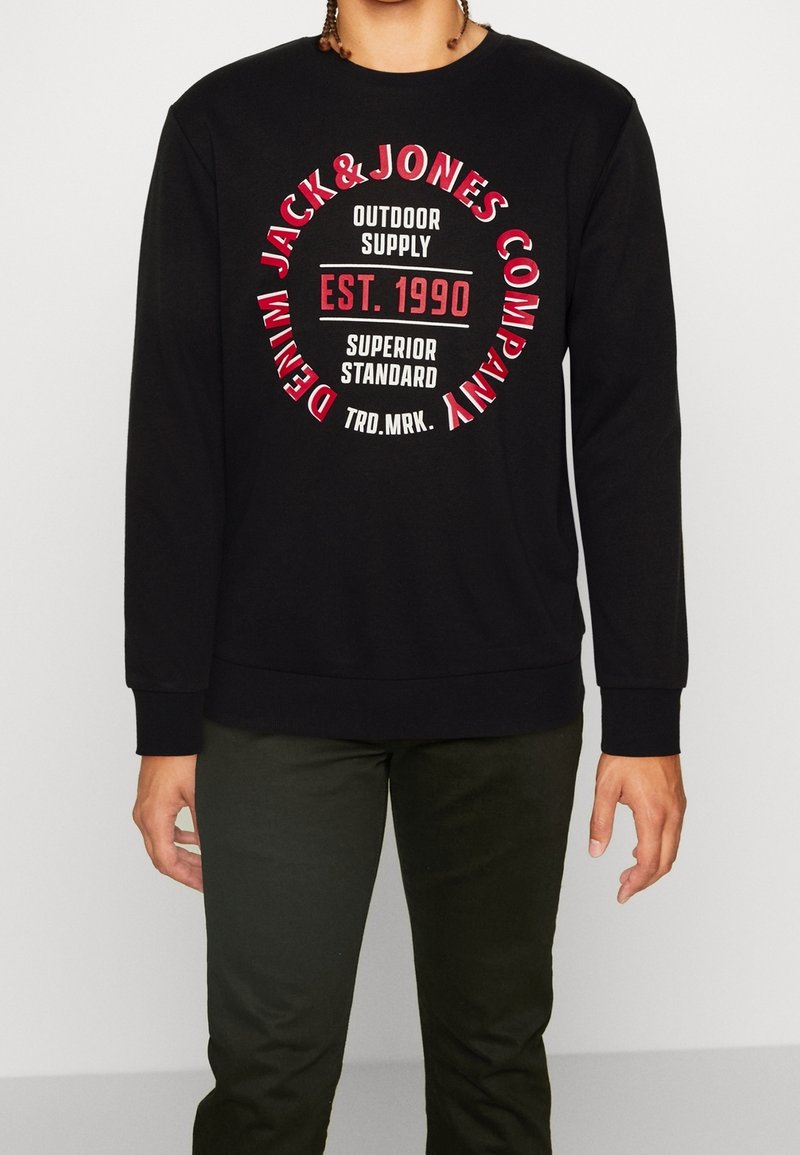 Sweat-shirt noir à manches longues avec texte circulaire rouge et blanc « Jack & Jones Company Denim » et « Est. 1990 Superior Standard » sur le devant.