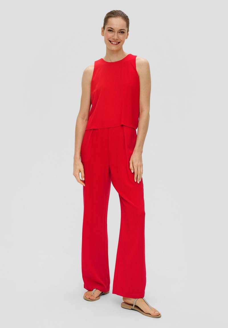 Roter ärmelloser Jumpsuit mit lockerer Passform, fließendem Stoff und weitem Bein. Kombiniert mit flachen Sandalen, die eine glatte Textur und ein klares Design zeigen.
