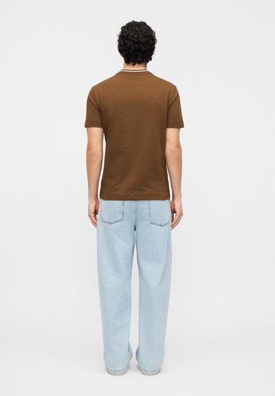 Άνδρας με σγουρά σκούρα μαλλιά που φοράει καφέ κοντομάνικο πουκάμισο και ανοιχτόχρωμα μπλε φαρδιά τζιν, στέκεται στραμμένος προς λευκό φόντο.