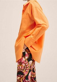 Chemise en coton orange avec une poche, coupe décontractée, design à boutons. Associée à un pantalon à motifs floraux roses, bruns et verts.
