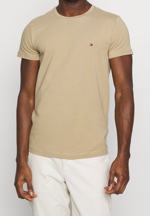 Mand iført beige T-shirt med korte ærmer og rund hals med lille logo på venstre bryst, kombineret med lyse bukser mod en ensfarvet baggrund.
