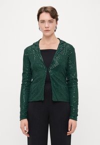 Cardigan vert à sequins avec capuche, présentant une coupe ajustée et des manches longues. Associé à un haut noir et un pantalon noir à jambes larges.