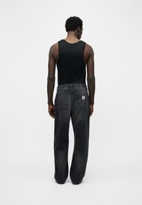 Άνδρας που στέκεται γυρισμένος, φοράει μαύρο αμάνικο και χαλαρό ξεθωριασμένο μαύρο cargo παντελόνι με μεγάλες πίσω τσέπες.