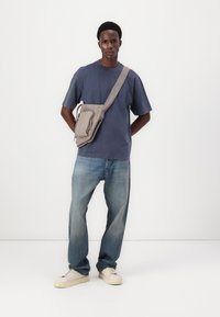 Siva crossbody torba s patentnim zatvaračem, nosi se preko tamnoplavog majice. Svijetloplave traperice, bijele tenisice. Jednostavan, funkcionalan dizajn.
