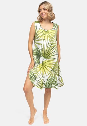 Uśmiechnięta kobieta w białej sukience bez rękawów z zielonym tropikalnym nadrukiem liści, stojąca boso na jednolitym tle.
