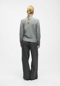 Leichter grauer Strickpullover mit Stehkragen, kombiniert mit weit geschnittenen schwarzen Hosen mit vertikalen weißen Streifen.