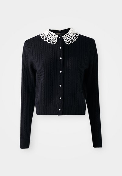 Knitted black cardigan featuring a decorative white lace collar, button closure, and ribbed texture. Long sleeves and cropped length.