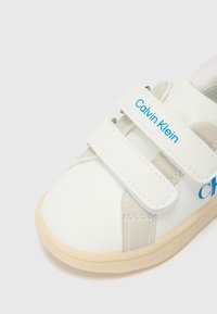 Vit sneaker med grå detaljer och blå "Calvin Klein"-logotyp. Har kardborrestängning och gummisulor. Slätt material, rundad form.