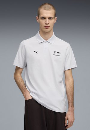 Giovane uomo con capelli corti, indossa una polo bianca BMW Motorsport con logo Puma, posa con una mano in tasca davanti a uno sfondo grigio.