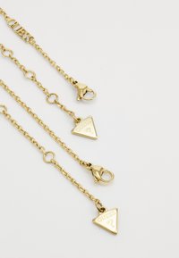 Accessoire de chaîne de couleur dorée avec des pendentifs triangulaires portant le logo Guess, deux fermoirs en homard et une finition lisse et brillante.