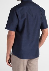 Chemise bleu marine à manches courtes en tissu texturé, avec un empiècement arrière droit et un petit détail de tag sur le côté.