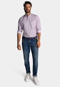 Lichtroze overhemden met kleine blauwe stippen, gecombineerd met blauwe jeans. Witte sneakers, opgerolde shirtmouwen en een leren bruine riem.