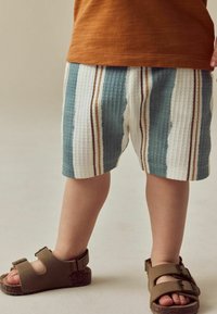 Gestreifte Shorts in Blau, Weiß und Brauntönen; aus leichtem, atmungsaktivem Material hergestellt. Kombiniert mit braunen Sandalen mit zwei Schnallen.