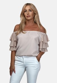 Jasnoróżowa bluzka typu off-the-shoulder z warstwowymi falbanami na rękawach, wykonana z miękkiego materiału, zestawiona z białymi dopasowanymi spodniami.