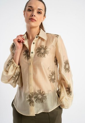 Kobieta w prześwitującej beżowej bluzce z rękawami balonowymi i wyhaftowanymi dużymi kwiatowymi wzorami, w zestawieniu z oliwkowozielonymi spodniami.