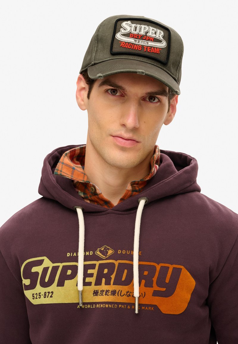 Gorra verde oliva con un parche rectangular que muestra "SUPER DRY RACING TEAM" en texto rojo y blanco; llevada con un hoodie morado que presenta un logo.
