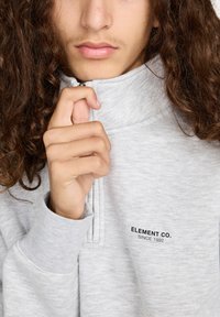 Persoon met lang krullend haar die een lichtgrijze Element Co. half rits sweatshirt met zichtbaar logo op de borst dichtritst.