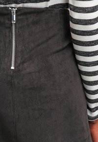 Jupe crayon en suède noir avec une fermeture éclair argentée visible à l'arrière, associée à un haut à manches longues noir et blanc à rayures. Texture douce.